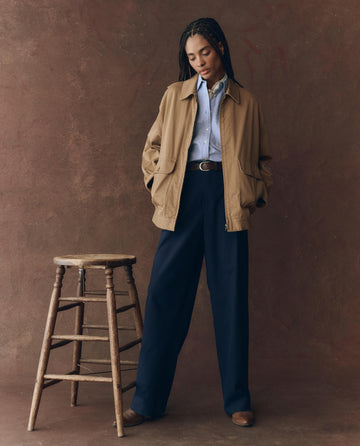 A person stands next to a wooden stool, wearing THE GREAT. F25 D1 The Folsom Jacket -- Tobacco over a blue shirt, navy wide-leg pants, and brown shoes, against a brown textured background.