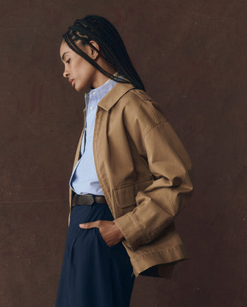 A person stands in profile against a brown background, wearing THE GREAT. F25 D1 The Folsom Jacket in Tobacco over a light blue shirt and dark trousers, with one hand in their pocket.