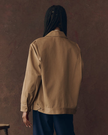 A person with braided hair, wearing THE GREAT. F25 D1 The Folsom Jacket in Tobacco and dark pants, stands facing away from the camera against a brown textured background.