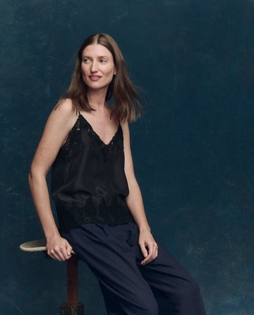 A woman with straight brown hair sits on a stool against a dark blue background, wearing THE GREAT. SP25 COLLECTORS The Legacy Top in black with adjustable straps and loose dark pants, looking slightly to her left.