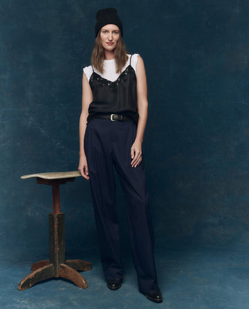 A woman stands by a wooden stool, layering THE GREAT. SP25 COLLECTORS The Legacy Top in black—featuring adjustable straps—over a white shirt, styled with pinstripe trousers and black shoes against a dark blue backdrop.