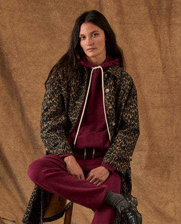 A person wearing a THE GREAT. H26 FLEECE BACK KNITS Fleece Pullover Hoodie in plum and matching sweatpants under a long leopard-print coat.