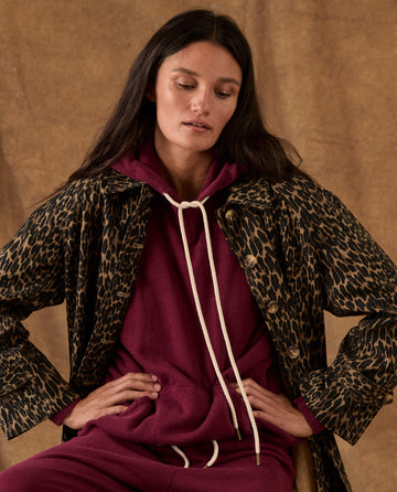 A person wearing The Fleece Pullover Hoodie. -- Plum, a dark plum fleece pullover with long white drawstrings, under a leopard-print button-down jacket.
