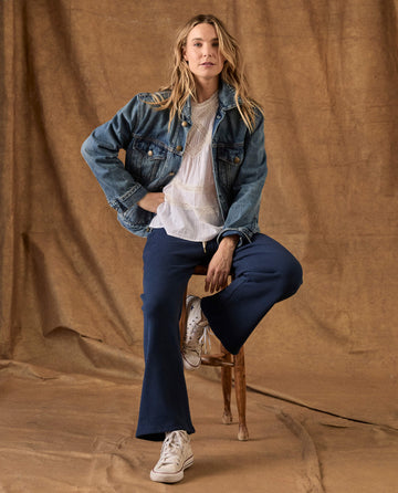 A woman wearing a denim jacket, white blouse, and THE GREAT. H26 FLEECE BACK KNITS The Fleece Lap Sweatpant in True Navy sits on a wooden stool.