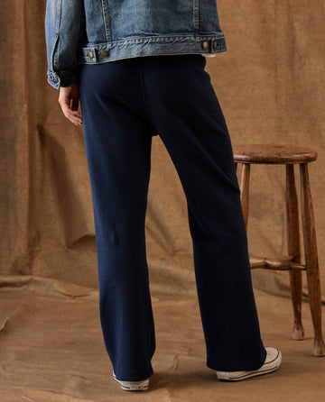 Back view of a person wearing True Navy The Fleece Lap Sweatpant, a denim jacket, and white sneakers, standing next to a wooden stool.