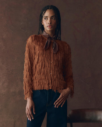 A woman stands indoors wearing THE GREAT. F25 D1 The Fringe Cardigan in bronze, paired with dark jeans and a neck scarf, with one hand resting on a chair.
