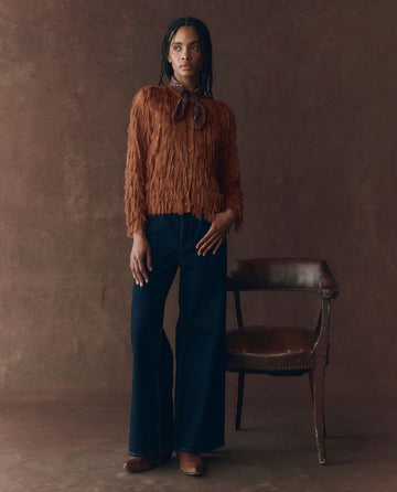 A woman stands beside a wooden chair wearing THE GREAT. F25 D1's The Fringe Cardigan in Bronze, paired with dark blue wide-leg jeans, brown shoes, and a neck scarf, set against a plain brown background.
