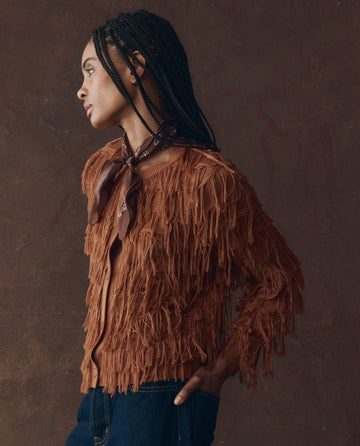 Woman in profile wears THE GREAT. F25 D1 The Fringe Cardigan in Bronze, a brown neck scarf, and dark blue jeans, standing against a textured brown background.