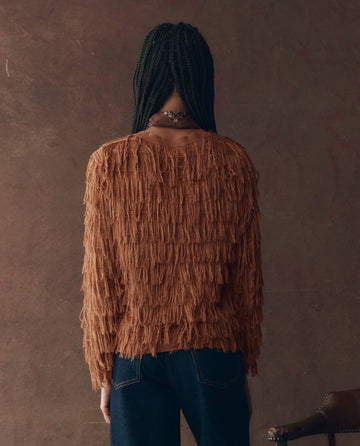 A person with long braids wears THE GREAT. F25 D1's The Fringe Cardigan in Bronze with dark jeans, standing with their back to the camera against a brown textured background.