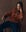 A person with braided hair sits in a wooden chair, wearing THE GREAT. F25 D1 The Fringe Cardigan in Bronze, styled with a brown scarf and dark jeans, looking directly at the camera.