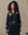 A woman stands against a beige background wearing THE GREAT. H25 COLLECTION's The Heirloom Rose Western Cardigan in black, paired with a black tiered skirt and belt.