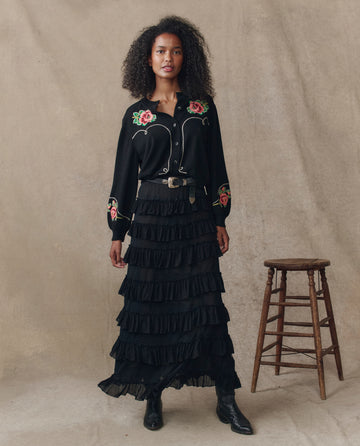 A woman indoors wears THE GREAT. H25 COLLECTION’s The Heirloom Rose Western Cardigan in black over a black floral-embroidered blouse, tiered skirt, and boots. A wooden stool stands beside her against a neutral backdrop.