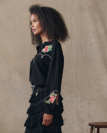 A woman with curly hair models THE GREAT. H25 COLLECTION's The Heirloom Rose Western Cardigan in black, featuring floral embroidery, styled with a tiered ruffle skirt, in profile against a plain background.