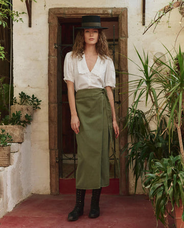 A woman stands before a gated doorway, wearing THE GREAT. PF25 The Manor Top in white, an olive skirt, black lace-up boots, and a black flat-brim hat, surrounded by potted plants.