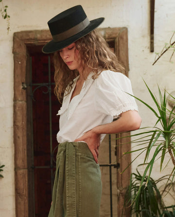 A person wears THE GREAT. PF25 The Manor Top in white with puff sleeves, a black wide-brimmed hat, and high-waisted green pants while standing indoors near a plant.