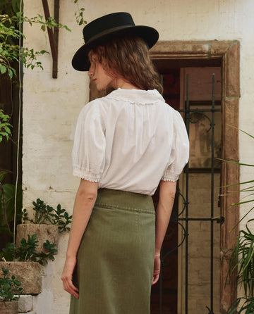 A person with wavy hair, wearing THE GREAT. PF25's The Manor Top in white and a green skirt, stands facing away in front of a rustic building while sporting a black wide-brimmed hat.