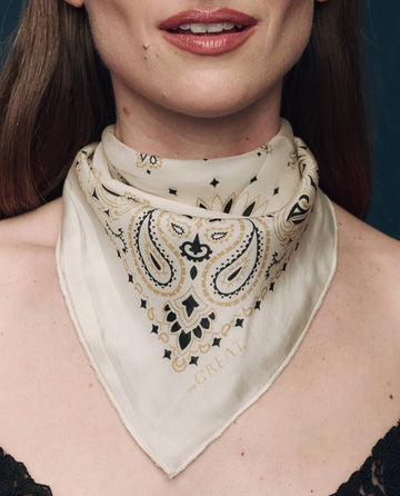 A person wearing The Silk Bandana in Cream by THE GREAT. F25 COLLECTORS, featuring a black and gold paisley pattern, tied around their neck in a close-up shot.