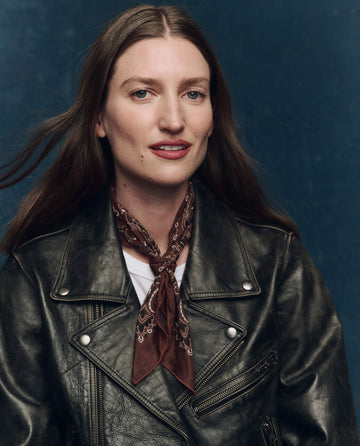 A woman with long brown hair wears a black leather jacket, white shirt, and THE GREAT. F25 COLLECTORS The Silk Bandana in Chocolate, posing against a dark blue background.