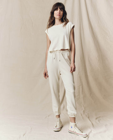 A person stands wearing THE GREAT. CORE KNITS' The Stadium Sweatpant in Washed White, a light beige casual outfit with short sleeves and jogger-style pants featuring an elasticated waist, paired with white high-top sneakers against a plain beige curtain backdrop.