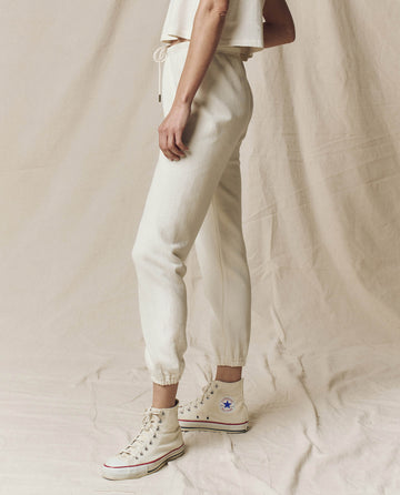 A person wearing The Stadium Sweatpant in Washed White by THE GREAT. CORE KNITS and high-top sneakers stands sideways against a beige background, evoking a vintage athletic vibe. Only the lower half of the body is visible.