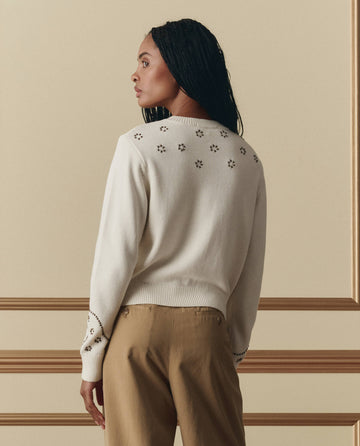 A woman with long braided hair faces away, wearing THE GREAT. SP26 D2 The Studded Cardigan in Natural and tan pants, standing against a beige wall with gold trim.