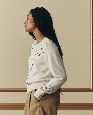A woman in profile wears THE GREAT. SP26 D2 The Studded Cardigan in natural, featuring small flower patterns, paired with tan pants and posed against a beige wall with decorative molding.