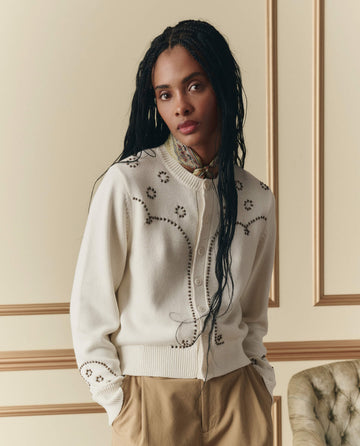 A woman with long braided hair models THE GREAT. SP26 D2 The Studded Cardigan in Natural and tan pants, standing indoors by an upholstered chair and a cream paneled wall.