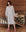 A person wearing The Sweatshirt Dress in Light Heather Grey by THE GREAT. HOL 24 SLEEP, paired with red socks, stands in a rustic wooden room next to a metal bed adorned with a patchwork quilt.