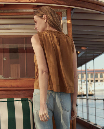 A woman wearing THE GREAT. SU26 The Vignette Top in Clay and light blue jeans stands on a boat deck, facing away from the camera.