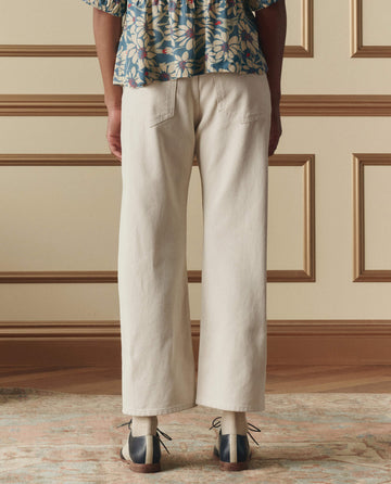 A person stands indoors facing away, wearing a blue floral top, THE GREAT. SP26 D2 The Studded Wayne Jean -- Natural, and two-tone shoes; the background features a wood-paneled wall and patterned rug.