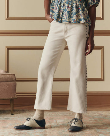 A person stands indoors wearing THE GREAT. The Studded Wayne Jean in Natural by SP26 D2, paired with a floral top, light socks, and black-and-white oxford shoes. Only the lower body is shown.