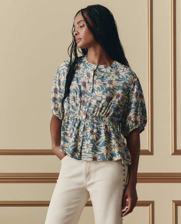 A woman stands before beige paneled walls wearing THE GREAT. SP26 D2 Hayflower Top in Blue Scattered Bloom, a puff-sleeve floral blouse, styled with cream pants.