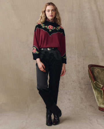 A woman stands against a beige backdrop wearing THE GREAT. H25 COLLECTION's The Heirloom Rose Western Pullover in Wine, paired with black pants and boots. An olive green chair appears partially on the right.