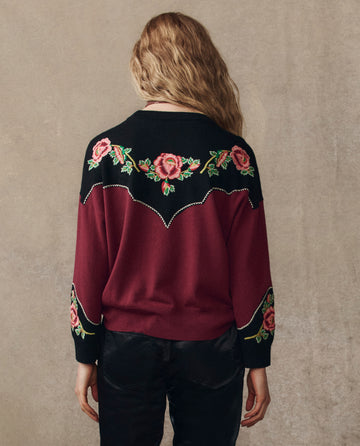 A woman with wavy hair faces away, wearing The Heirloom Rose Western Pullover in wine by THE GREAT. H25 COLLECTION, featuring maroon and black colors with floral embroidery on the shoulders and sleeves.