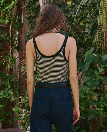 A person stands outdoors with their back to the camera, wearing THE GREAT. PF25 The Henley Slim Tank in Black & Cream Stripe, blue jeans, and a scarf, surrounded by greenery.
