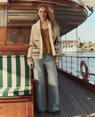 A woman stands on the deck of a sailboat wearing THE GREAT. SU26 The Sailboat Lodge Cardigan in cream, paired with a brown top and wide-leg jeans. Water and buildings appear in the background.