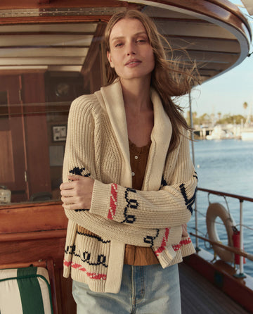 A woman in THE GREAT. SU26 The Sailboat Lodge Cardigan – Cream and jeans stands on a boat with the marina and water behind her.
