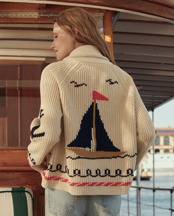 A woman stands on a boat in THE GREAT. SU26 The Sailboat Lodge Cardigan in cream, featuring wave and seagull motifs on the back, with a harbor visible behind her.