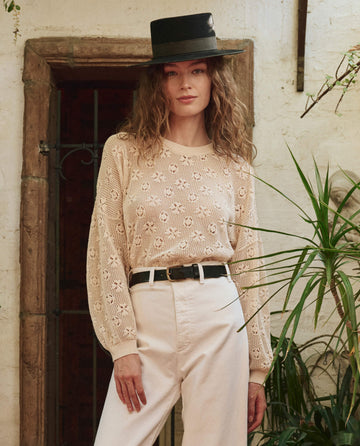 A woman stands indoors near a plant and a decorative doorway, wearing THE GREAT. PF25's The Crochet Pullover in Natural, paired with a wide-brimmed black hat and high-waisted light pants.