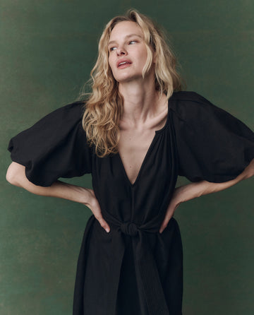 A woman with wavy blonde hair stands against a green background, hands on hips and looking upward, wearing THE GREAT.’s The Fairground Dress in Black.