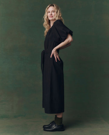 A woman stands in profile against a green backdrop, wearing THE GREAT. F25 D2’s The Fairground Dress in black with puffed sleeves, paired with dark socks and black shoes.