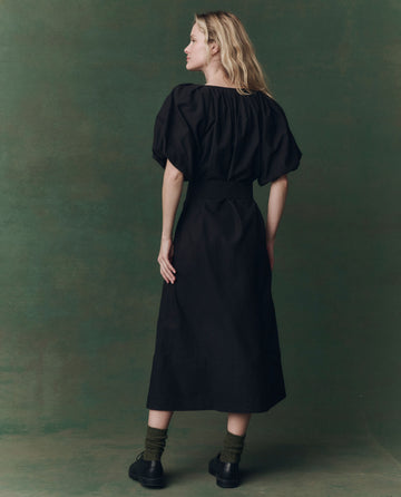 A woman stands with her back to the camera, wearing THE GREAT. F25 D2 The Fairground Dress in black with puffed sleeves, paired with green socks and black shoes against a green background.