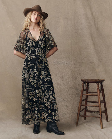 A woman wearing THE GREAT. H25 COLLECTION's The Festal Dress – Brocade Floral stands beside a wooden stool against a neutral fabric backdrop, accessorized with a wide-brimmed hat and black boots.