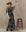 A woman in a brown hat and The Festal Dress -- Brocade Floral by THE GREAT. H25 COLLECTION stands beside a wooden stool against a plain beige backdrop.