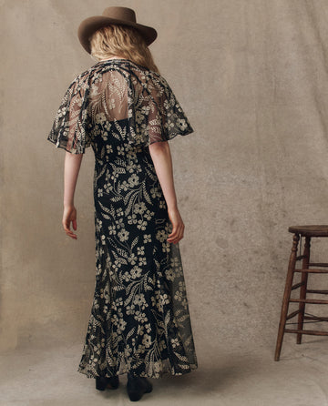 A woman faces away, wearing THE GREAT. H25 COLLECTION’s The Festal Dress in Brocade Floral—black with sheer embroidered overlay—paired with a brown wide-brim hat and black shoes. A wooden stool stands at her right.