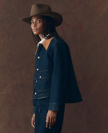 A person wearing THE GREAT. F25 D1 The Denim Swingy Jacket – Midnight Rinse stands against a brown textured background.