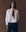 A person stands against a brown background wearing THE GREAT. F25 D1 The Cropped Oxford Shirt in white, dark jeans, and a brown necktie, with one hand in their pocket.