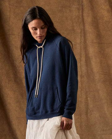 A person wearing The Fleece Pullover Hoodie in True Navy, featuring a navy blue pullover design with thick, knotted drawstrings and a front pouch pocket.
