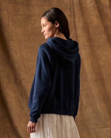 A person wearing a THE GREAT. H26 FLEECE BACK KNITS The Fleece Pullover Hoodie in true navy over a white skirt.