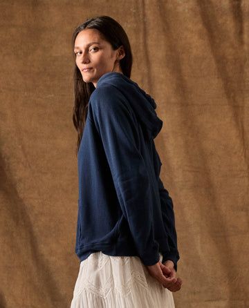 A person wearing a THE GREAT. H26 FLEECE BACK KNITS The Fleece Pullover Hoodie in True Navy over a white skirt, looking over their shoulder toward the camera.